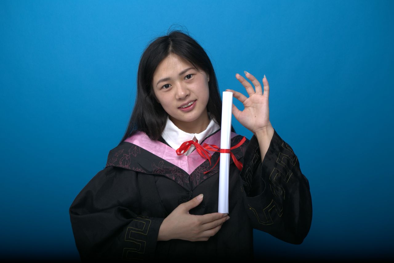 Asian female graduate holds diploma against blue background, smiling.