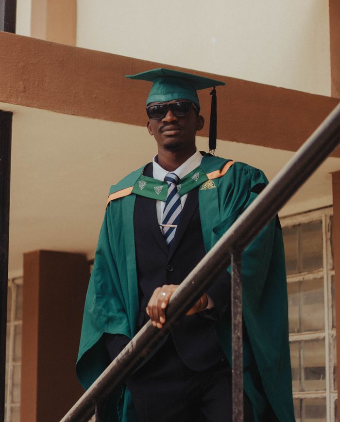 Graduation portrait of a confident young man in green cap and gown standing outdoors.