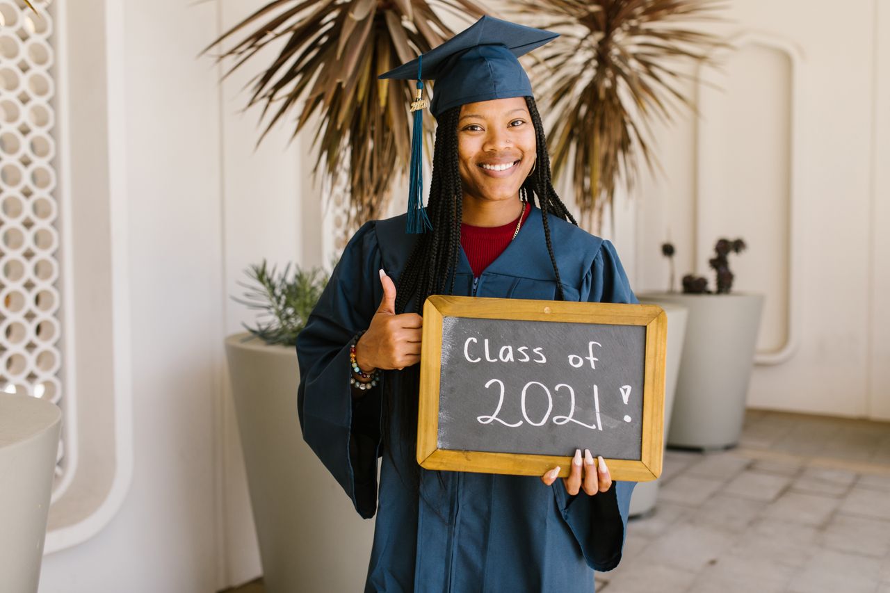 A smiling graduate holding a Class of 2021 sign with a thumbs up gesture.